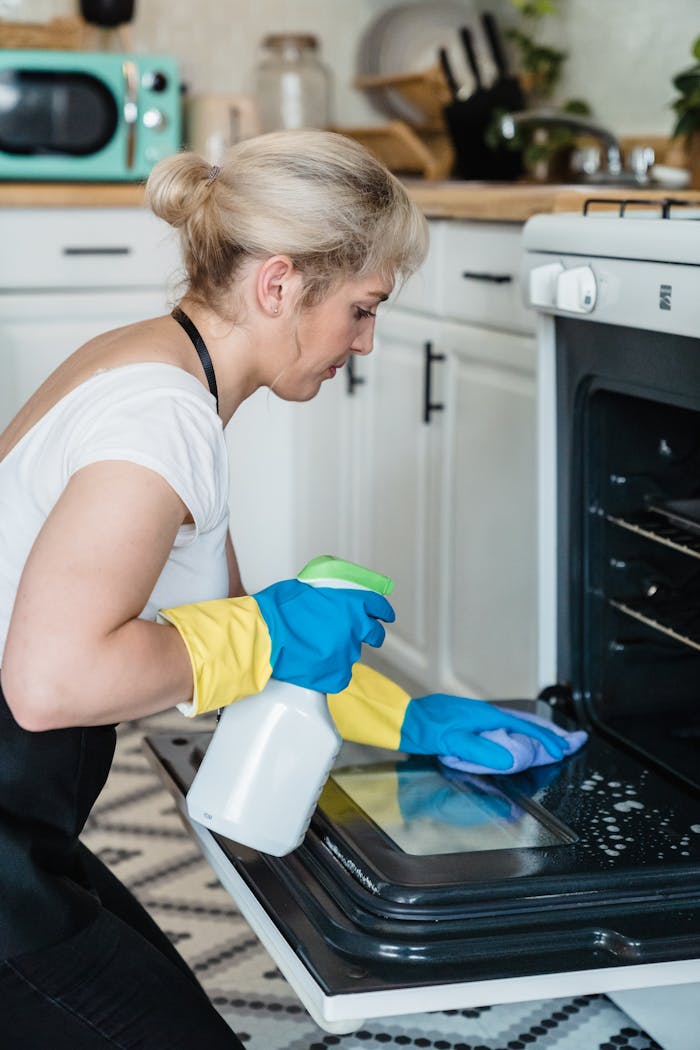 A woman uses a spray bottle and cloth to clean an oven in a modern kitchen setting.